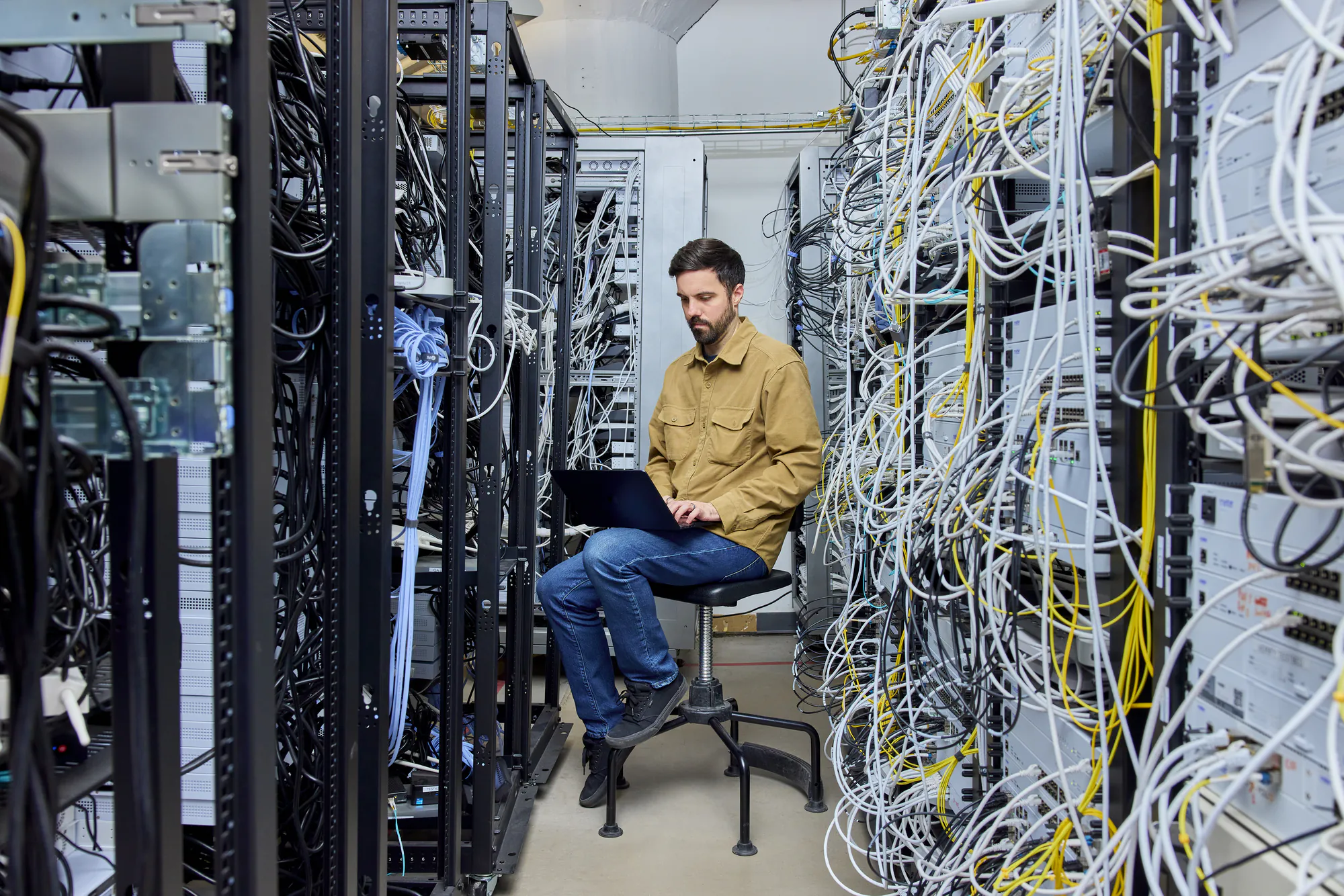 Engineer on a laptop between rows of cabled network racks in a data center